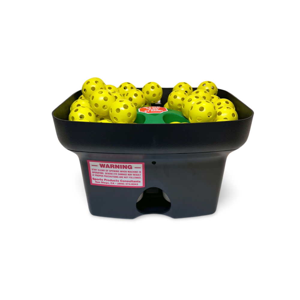 Pitching machine from the front, hopper full of yellow small balls, warning label on the housing